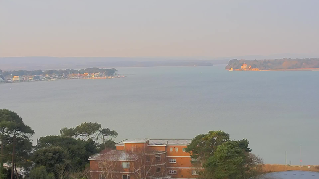 A scenic view of a calm body of water with gentle ripples, reflecting soft light under a clear sky. To the left, a marina is visible with a row of sailboats and nearby buildings. In the foreground, a tree line with evergreen trees can be seen, and in the lower right corner, a portion of a brown building is partially visible. The background features rolling hills fading into the horizon, creating a peaceful and serene landscape.