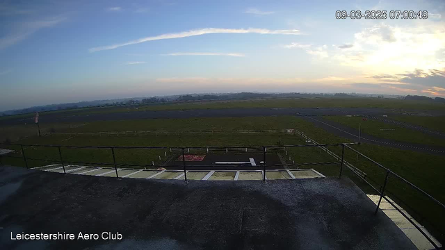 A view from a rooftop terrace showing a large, open grassy field with a runway. The sky is light blue with some wispy clouds, indicating early morning. In the foreground, there is a railing along the edge of the terrace, and a small portion of the roof is visible. The runway is marked with white lines, and there are some signs and fences around the area. The setting appears calm and serene, with no visible activity.