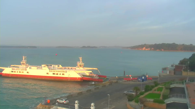 A calm waterfront scene featuring a large ferry docked at a pier with a red and white color scheme. The water is blue and rippling, with a few distant islands visible in the background. The sky is light with some clouds, suggesting early morning or late afternoon. On the shore, there are vehicles parked and a small building with signs. Green shrubs and a landscaped area are situated near the edge of the dock.