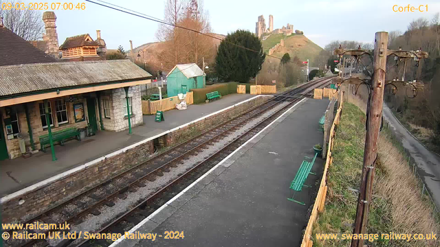 A view of a railway station with a platform and surrounding area. The station has a stone façade, a sloped roof, and green benches. To the left, a wooden building stands with some greenery nearby. The platform extends along the tracks, which are visible in the foreground. In the background, a hill features a castle ruin. The scene is set in early morning light, with a clear sky, and there is a wooden fence along the pathway beside the tracks.