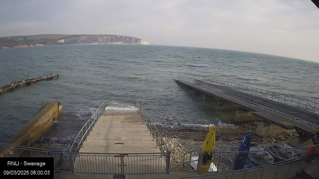 A view of a calm sea with gentle waves, taken from a pier. In the foreground, there are concrete steps leading down to the water, surrounded by a metal railing. On the right, there are two docks extending into the water, with a yellow kayak and a blue kayak resting on the shore. In the background, there are rocky cliffs and rolling hills under a cloudy sky. The image is timestamped 09/03/2025 at 08:00:03.