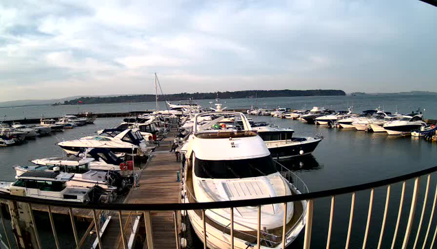 A view of a marina with numerous boats lined up along wooden docks. Several white and blue yachts are anchored in the water, reflecting the cloudy sky above. In the background, there is a distant shoreline with trees, and the atmosphere appears calm and serene. A metal railing is visible in the foreground, framing the scene.