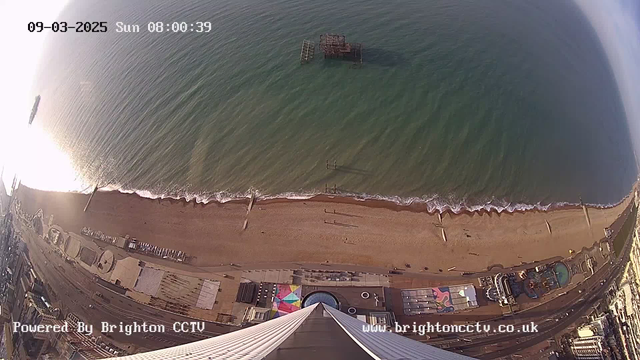 A high-angle view of a beach and sea, taken from a webcam. The scene captures a sandy beach with several long, narrow objects likely to be beach huts or sun loungers lined up. Waves gently lap at the shore. To the left, a pier extends into the water. In the background, the sea is a deep blue, reflecting sunlight, and a few people are seen walking along the beach. There are some amusement rides and structures near the edge of the beach. The date and time are displayed in the top left corner, and a watermark appears in the bottom left.