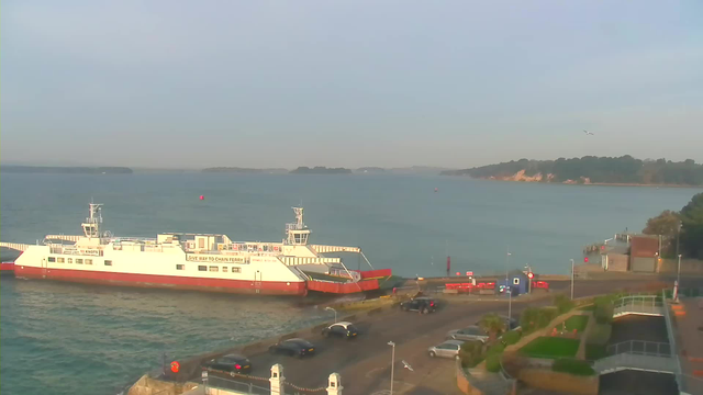 A white and red ferry is docked at a waterfront with calm blue waters. The ferry is positioned at a pier with a small ramp extending towards it. In the background, there are green hills and trees lining the shore. Several cars are parked along the waterfront road, and there are some signs and red barriers near the pier. The sky is mostly clear with a few clouds.