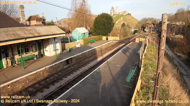 The image shows a railway station platform with a view of train tracks leading away. On the left, there is a stone building with large windows, and a green bench is visible on the platform. In the background, there are green trees and a wooden fence. On a hill in the distance, the ruins of a castle can be seen. The sky is clear, indicating a sunny day.