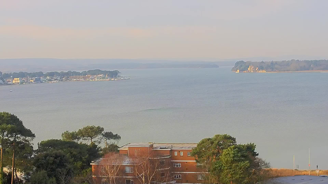 A serene view of a calm body of water with a distant shoreline. On the left, a marina can be seen with several small boats and buildings, while the right side reveals an island featuring a structure resembling a castle or historic building, surrounded by trees. In the foreground, there are some trees and a low, brown-bricked building, adding depth to the scene. The sky is light and cloudy, suggesting a tranquil atmosphere.