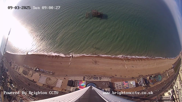 Aerial view of a sandy beach beside the sea. The water is calm with gentle waves lapping at the shore. In the distance, there is a weathered pier jutting out into the ocean. Scattered people walk along the beach. The scene also includes colorful beach structures and a boardwalk area, while a tall building or observation tower reaches upward from the bottom of the image. The sky is clear with soft sunlight reflecting off the water, creating a bright sheen on the surface.