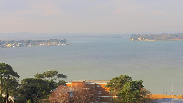 A scenic waterfront view showing a calm body of water with a soft gradient of colors ranging from blue to green. In the background, there are trees and buildings along the shoreline, with several boats visible on the water. On the right side, there’s a small island featuring a structure that may be a lighthouse or a historical building, surrounded by trees. The foreground includes a mix of greenery and a low-rise building with an orange facade. The sky is hazy, suggesting a tranquil atmosphere.