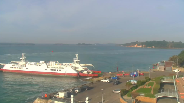 A white and red ferry is docked at a harbor, with the water gently rippling around it. The ferry has a sign indicating "Give way to chain ferry." There are several parked cars in the foreground, including a white van and a blue car. In the background, there is a tree-covered shoreline and a few small boats moored at a distant pier. The sky is clear with a few soft clouds, and the scene is illuminated by sunlight.