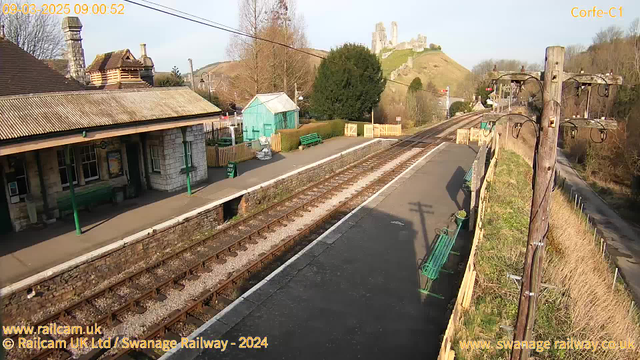 A view of a railway station in Corfe, England, featuring a platform with benches made of green metal. In the background, there is a stone building with a sloped roof and several trees. A light green shed is visible to the left. The area is surrounded by rolling hills, with a castle ruin perched on a hilltop in the distance. The railway tracks extend along the platform, and there are wooden fences and signs indicating exits nearby. The scene is bathed in bright sunlight on a clear day.