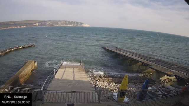 A view of a coastline featuring a calm sea under a partly cloudy sky. In the foreground, there is a concrete ramp leading down to the water, bordered by a metal railing. To the left, a wooden pier extends into the sea. Several colorful kayaks are lined up on the shore, with a collection of rocks visible beneath the water. In the distance, cliffs rise along the coastline. The scene suggests a tranquil maritime environment.