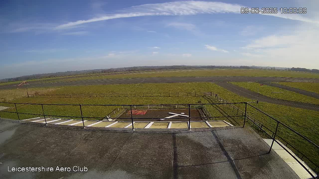 A wide-angle view of an open landscape from a webcam at the Leicestershire Aero Club. The foreground shows a railing with a flat, paved area. Below the railing is a grassy field with a marked helipad featuring a white X shape. In the distance, there is a runway and sections of mown grass, while scattered patches of blue sky and a few clouds above create a clear, sunny atmosphere.