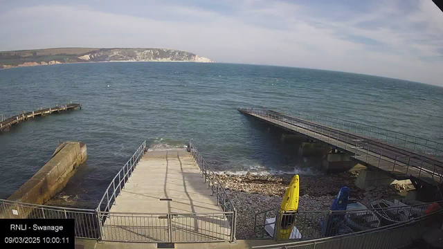 A coastal scene featuring a clear blue sea with gentle waves. In the foreground, a wide concrete ramp leads down to the water, bordered by a metal railing. On the right side, there are two parked kayaks, one yellow and one blue. To the left, a wooden jetty extends into the water, curving out of view. In the background, a rocky cliff rises above the shoreline, partially covered with greenery under a bright sky. The image is timestamped with "09/03/2025 10:00:11."