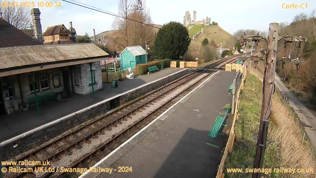 A railway station scene with a platform featuring multiple green benches and a stone building. In the background, a hill topped with a castle ruins is visible. A wooden pole with electrical wires stands on the right side of the image. The sky is clear with a few clouds, and the overall setting appears peaceful, with trees and a wooden fence surrounding the area.