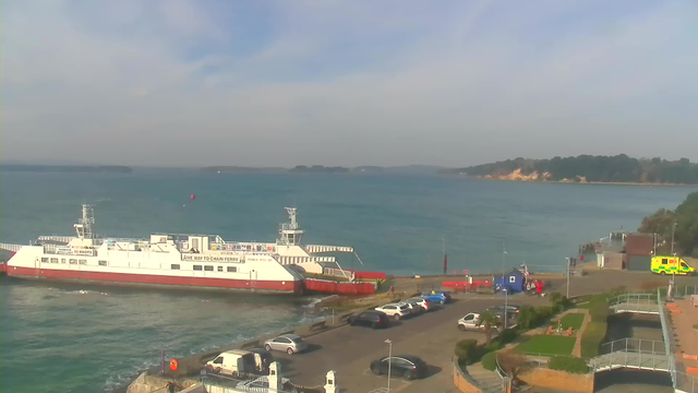 A large white ferry with red accents is docked at a harbor, facing the water. The sea is calm with a light blue hue, and several small boats can be seen in the distance. In the foreground, there are parked cars along a paved area, and a green ambulance is visible near a small blue structure. To the right, there are landscaped gardens with seating areas, and a walkway leads towards the waterfront. The coastline with trees and cliffs can be seen in the background under a partly cloudy sky.