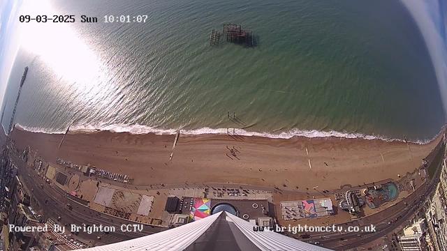 Aerial view of a sandy beach with gentle waves lapping at the shore. In the foreground, there are colorful umbrellas and structures, possibly beach facilities. People can be seen walking along the beach. Off to the side, a pier extends into the water, and a dilapidated structure is visible in the ocean. The sky is clear with some wispy clouds, and the sun is reflecting off the water, creating a bright patch on the surface. The image is timestamped with the date and time in the upper left corner.