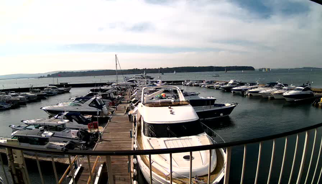 A view of a marina filled with various boats docked at wooden piers. In the foreground, a large white yacht is prominent, with several smaller boats surrounding it. The water is calm, reflecting the sky, which is partly cloudy. In the background, distant land is visible, along with additional boats and sails on the water, indicating a recreational atmosphere. The image captures a sunny day at the marina.