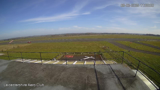 A view from a webcam positioned on a building at an aerodrome. The foreground shows a flat rooftop with a railing, and in the distance, there is a vast expanse of green grass and an airstrip. A small red aircraft is visible on the runway, which is black with white markings. The sky above is mostly clear with a few wispy clouds. 