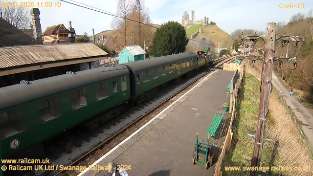 The image shows a railway station with a green passenger train parked on the tracks. The scene features a platform made of grey concrete, bordered by grass and wooden fencing. In the background, there are trees and a hill topped with ruins of a castle, partially obscured by bushes. A wooden building with a sloped roof is visible on the left side of the image. Several people can be seen on the platform, and a person is walking along a path in the distance. The sky is clear with some soft sunlight.