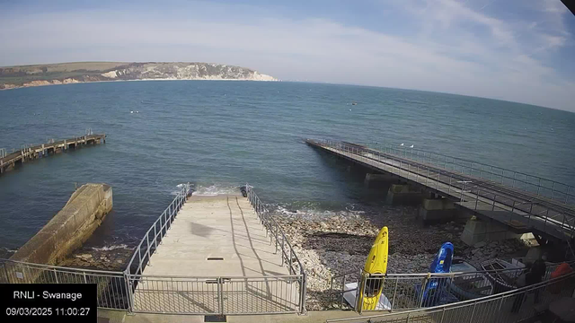 A view of the coastline featuring calm blue waters and a white rocky cliff in the background. In the foreground, there is a concrete ramp leading to the water, bordered by metal railings. To the right, two colorful kayaks, one yellow and one blue, are secured on the shore. A wooden jetty curves into the water from the left side of the image, while a larger jetty extends straight ahead. The sky is clear with a few wispy clouds.