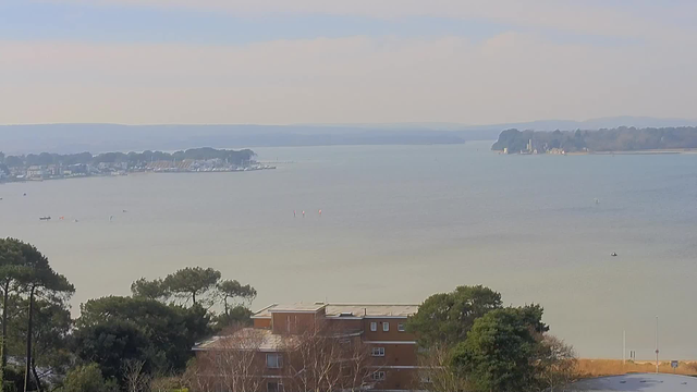 A view of a calm body of water with a soft, muted sky above. On the left side, a line of buildings can be seen along the shore, including houses and a marina with boats. Several small craft are visible on the water, along with floating markers. In the distance on the right, a tree-lined island emerges from the water, featuring structures, likely buildings, partially obscured by trees. The foreground includes low greenery and some trees, with a residential building located close to the shore. The overall scene conveys a serene, coastal atmosphere.