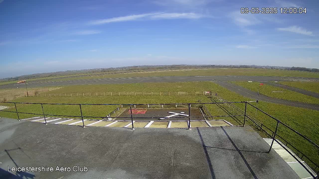 A view from a webcam at Leicestershire Aero Club showing a grassy airfield. In the foreground, there is a black landing area marked with a white crossed pattern. In the background, there are runways and patches of grass, with a clear blue sky above. Fencing runs along the edges of the airfield, and a small sign is visible on the right side of the image. The scene appears to be bright and sunny.