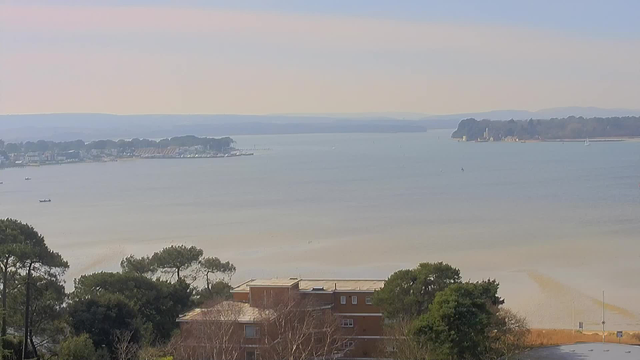 A scenic view of a calm body of water, with land on both the left and right sides. In the foreground, there are several trees and a brick building. The water appears still, reflecting a soft blue sky, while distant hills are visible on the horizon. A few small boats are scattered across the water. The overall atmosphere is peaceful and serene.