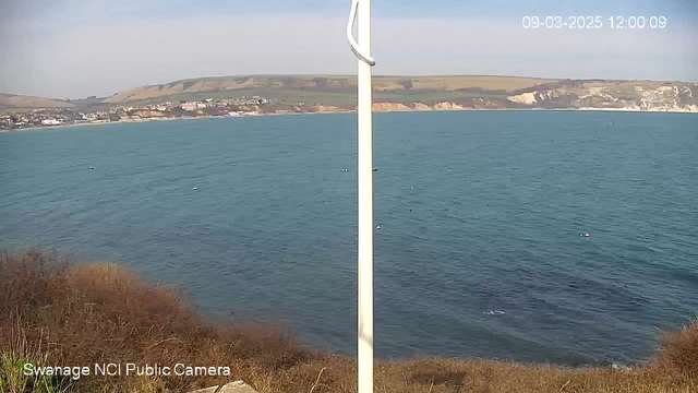 A view of the sea with gentle waves, showcasing a calm blue water surface. In the background, a coastal town can be seen with houses nestled against green hills, leading up to rocky cliffs on the right side of the image. The foreground contains some low shrubs and grass, and a white pole stands upright, slightly right of center. The sky is clear with a few clouds.