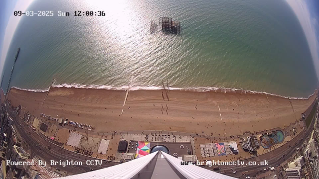 Aerial view of a sandy beach with gentle waves and people walking along the shore. A pier extends into the water from the left side of the image. Below, there are various colorful structures and attractions along the beachfront, including a circular building and a rectangular painted area. The sun is shining, creating a bright reflection on the water. The sky is mostly clear with some light clouds.