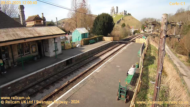 A sunny railway station scene featuring a stone platform with a couple of green benches. In the foreground, a person sits on one of the benches, facing the tracks. To the left, there is a station building with a peaked roof and a sign. A small green shed is situated nearby. The background showcases a hill with a castle ruins visible at the top. Alongside the tracks, a wooden utility pole with overhead wires can be seen. The surroundings include some trees and hedges. The sky is clear with minimal cloud cover.