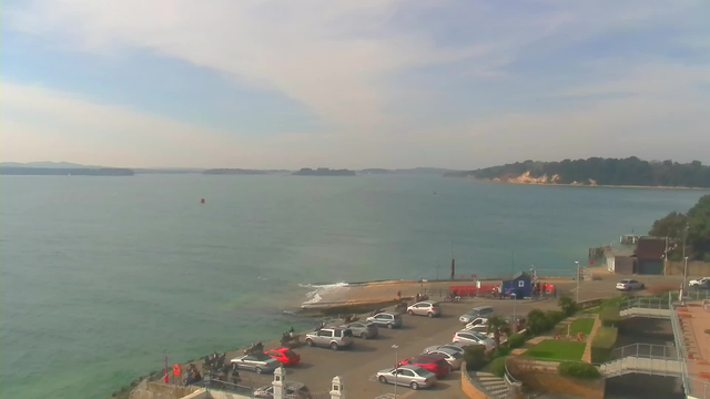 A scenic view of the coastline featuring calm blue water under a light blue sky with wispy clouds. In the foreground, there is a parking area filled with various cars, including red, gray, and white vehicles. A few people are gathered near the water's edge. To the right, a small blue structure and a red area are visible. In the distance, there are islands and a faint outline of a coastline on the horizon. The overall atmosphere is serene and captures a sunny day by the water.