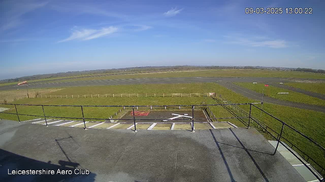 A clear blue sky with a few wispy clouds overhead. Below, a green grass landscape stretches out, with a large, open area resembling a runway. A red windsock is visible on the left side, indicating wind direction. In the foreground, there is a helipad marked with a white "X" surrounded by a railing, and the area is bordered by a wooden fence. The scene conveys a tranquil, sunny day at an airfield.