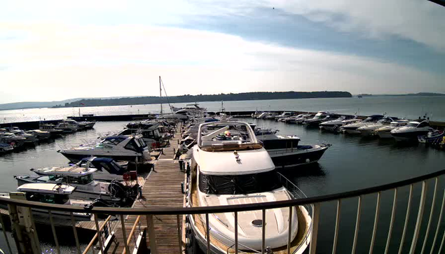 A waterfront scene featuring a marina filled with numerous boats. In the foreground, a large white yacht is docked, with other smaller boats around it. The background shows a calm body of water stretching towards a distant shoreline and a clear sky. The image captures the wooden dock leading to the boats, with a few masts visible from boats further out.