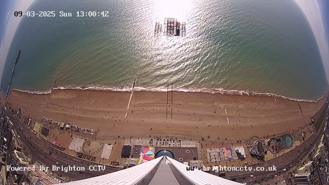 Aerial view of a beach with golden sand and ocean waves. The water reflects sunlight, creating a shimmering effect. In the distance, a pier extends into the sea, and several people are walking along the beach. The bottom of the image shows a colorful area with attractions, including a circular structure and various pathways. The scene conveys a sunny day at the seaside, with clear skies and minimal clouds.