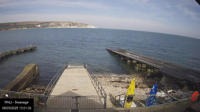 A view of a seaside area featuring a calm blue sea under a clear sky. In the foreground, there is a wooden pier extending into the water on the right, and a concrete ramp leading to the shore on the left, bordered by a railing. To the right of the ramp, there are several kayaks stored on a rocky beach, including yellow and blue ones. The background shows a white cliff along the coastline with green hills behind. The date and time are displayed at the bottom left corner.