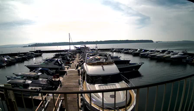 A marina scene featuring numerous boats docked in calm water. In the foreground, a white yacht is prominently visible, with other boats of various sizes lining the docks. The background shows a vast body of water under a partially cloudy sky, with a distant shoreline visible. The image conveys a peaceful, sunny day at the harbor.