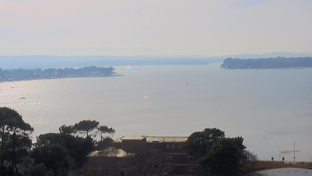 A wide view of a calm body of water under a pale sky. Sunlight reflects off the surface of the water, creating a shimmering effect. In the distance, there is a faint rainbow visible on the horizon, indicating a spot where sunlight interacts with moisture. On the left, a shoreline features trees and small buildings, while the right side shows more land and a few people walking along a path. The overall scene conveys a serene landscape with a soft color palette.