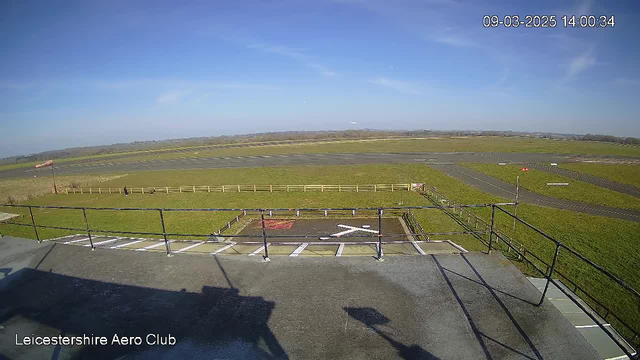 A clear blue sky extends overhead, with a few wispy clouds scattered throughout. Below, a wide grassy field is visible, extending towards a tarmac runway that runs horizontally across the center of the image. To the left, a small windsock is positioned near the edge of the field. A prominent helicopter landing pad marked with an 'X' is located in the foreground. A fence outlines part of the field, and there are a few scattered figures in the distance, likely engaging in recreational activities.