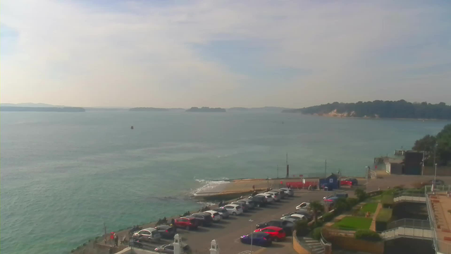 A tranquil seaside view featuring calm turquoise water under a bright sky scattered with clouds. In the foreground, a parking lot is filled with various cars, including red, white, and black vehicles. To the right, there's a well-maintained area with greenery and curving pathways. In the background, small islands and trees outline the horizon, while a distant sandy shore is visible along the coastline.