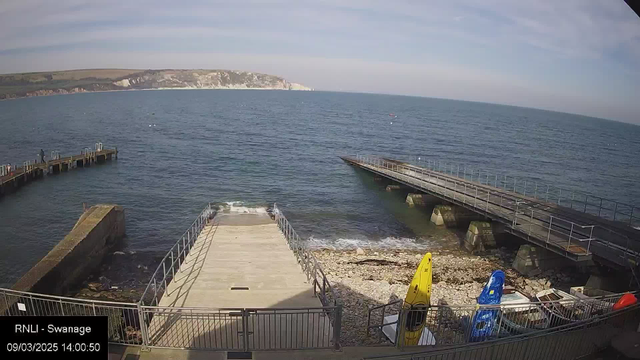 A coastal scene showing a calm sea under a mostly clear sky. In the foreground, a stone and concrete ramp leads down to the water, bordered by metal railings. On the left, a wooden pier extends into the water, with a person standing at its edge. To the right, there is another pier that also leads out over the water. Nearby, two brightly colored kayaks, one yellow and one blue, are positioned on a rocky shore, with some water lapping at the rocks below. In the distance, the coastline is visible, with cliffs and greenery rising above the waterline.