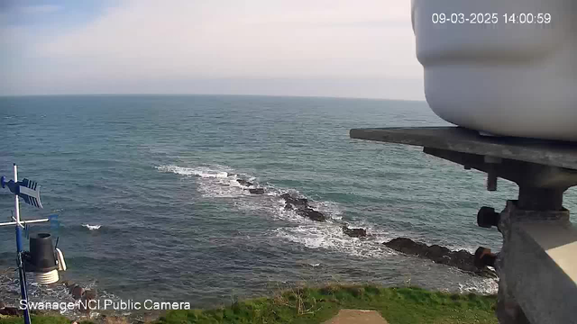 A view of the ocean on a clear day, with gentle waves lapping against a rocky coastline. The horizon is visible where the water meets the sky, which is mostly clear with some clouds. In the foreground, there is a metal structure with a weather vane and a light, along with a white container positioned on a flat surface. The scene conveys a peaceful coastal atmosphere.