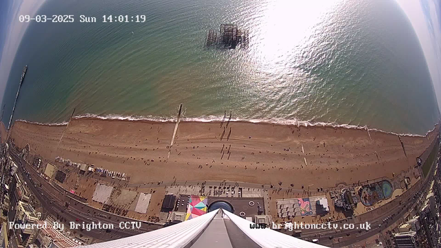 A bird's-eye view of a sandy beach with people walking along the shoreline. The water is calm and sparkles in the sunlight. A pier extends into the water, with a structure visible at the end. In the foreground, colorful beach huts and a funfair area can be seen, along with some people gathering. The sky is mostly clear with some light clouds. The date and time display shows "09-03-2025 Sun 14:01:19" at the top.