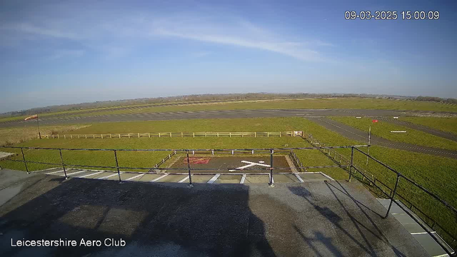 A clear blue sky stretches across the scene, with a few wispy clouds. Below, a vast green field is visible, bordered by a wooden fence. In the foreground, there is a flat rooftop with a rail. An airstrip is seen in the distance, marked with white lines on the tarmac. A windsock stands tall, indicating wind direction. The overall atmosphere is calm and open.