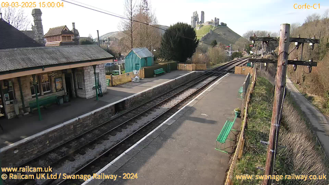 The image shows a railway station with a stone platform and several green benches. In the background, there is a hill with a castle ruins visible at the top. The scene is set on a clear day with trees and a wooden fence around the station area. There are power lines above, and the railway tracks run parallel to the platform. A small green shed is situated near the platform, and there are some railings along the edge of the platform.