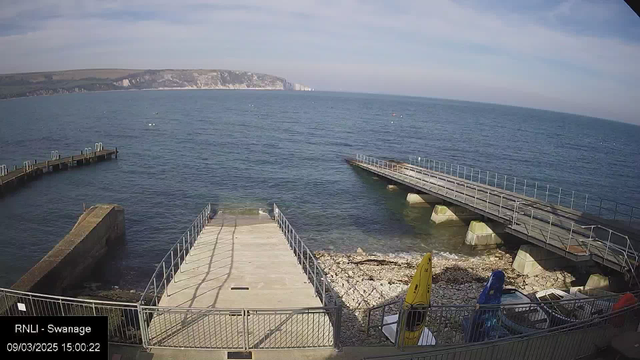 A view of a coastline with calm blue water under a partially cloudy sky. In the foreground, there is a concrete ramp leading down to the water, bordered by a metal railing. To the left, a wooden pier extends over the water with several mooring posts visible. On the right, a second pier extends out, also with railing. The shore is rocky, with some visible seaweed. In the lower right corner, there are two kayaks, one yellow and one blue, along with some other equipment. The date and time are displayed at the bottom left.