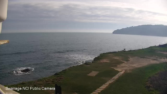 A view of the ocean under a slightly cloudy sky. The water appears calm with gentle waves lapping against the shore. In the foreground, there is green grass and a patch of sandy ground with a few stone path markers leading towards the ocean. On the right side, a grassy cliff rises, meeting the horizon.