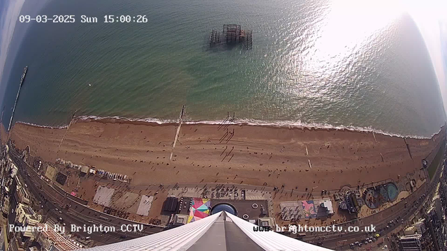 A high-angle view of a beach scene on a sunny day. The golden sand stretches along the shoreline with small groups of people scattered on the beach. In the water, a few small boats are visible. In the background, a pier extends into the sea, with a partially submerged structure nearby. There is a colorful beachside area with buildings and attractions, including a swimming pool and rides, below the viewpoint. The sky is clear with a few clouds, and the sunlight creates a shimmering effect on the water.