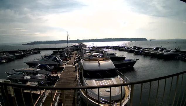 A panoramic view of a marina with numerous boats docked along a wooden pier. Several large yachts and smaller boats are visible, reflecting light from the sun. The water appears calm, with a horizon line of land in the distance and a cloudy sky overhead. The scene conveys a peaceful, early afternoon atmosphere.