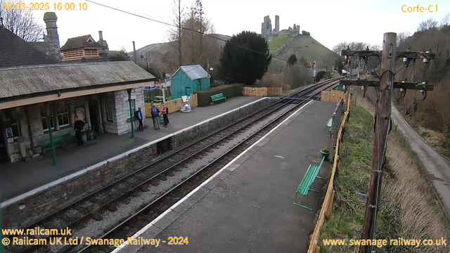 A railway station platform is visible with several green benches. A small group of people are standing near the station building, which has a stone facade and a roof with tiles. In the background, there is a green hill with a castle-like structure at the top. The area is surrounded by trees and a wooden fence, with railway tracks running along the platform. A telephone pole is on the right side of the image, with wires running overhead. The sky appears slightly overcast.