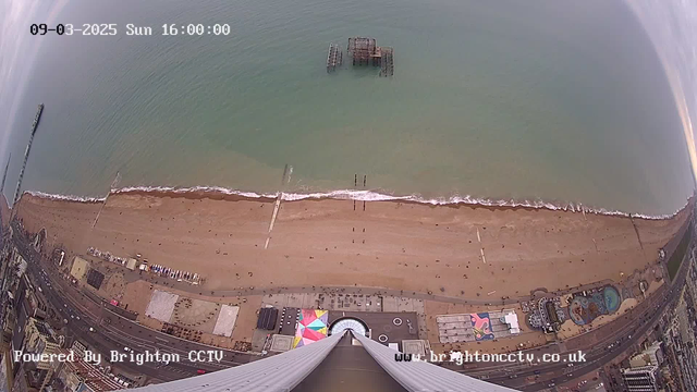 Aerial view of a beach with a wide sandy shoreline and gentle waves lapping the coast. There are people walking along the beach and several piers visible in the distance. An old, partially submerged pier structure is outlined in the water. The lower part of the image shows a promenade with colorful structures and attractions, including a circular building at the center. The sky is partly cloudy, and the scene captures a serene coastal environment.